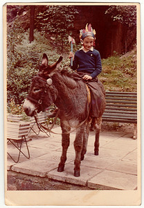 复古照片显示男孩坐在驴子上，他戴着印第安头巾并拿着战斧。