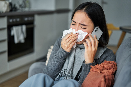 温度和人摄影照片_生病的韩国年轻女性生病、打喷嚏和拿着餐巾纸、生病待在家里、感冒了的画像。
