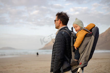 日落时分，父亲在西班牙兰萨罗特岛法马拉多风的沙滩上，背着他的婴儿男孩儿子在背包里享受纯净的大自然。