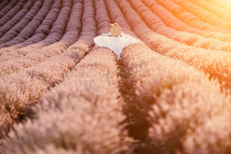 紫色氛围摄影照片_穿着白色连衣裙和草帽的快乐女人在日出时漫步穿过薰衣草田，沉浸在宁静的氛围中。