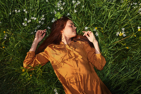 躺着休息摄影照片_一个放松的女人，躺在绿草地上休息，穿着橙色长裙，闭着眼睛，脸上挂着愉快的微笑，在夏日夕阳的温暖光线照耀下恢复体力