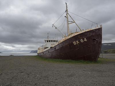 旧的废弃生锈渔船残骸站在冰岛西峡湾的沙滩海岸、戏剧性的灰色天空和悬崖背景