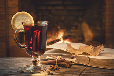 葡萄酒摄影照片_在带燃烧壁炉的舒适房间里，桌上放着一杯热葡萄酒、香料和一本书