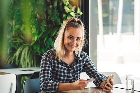 微笑的金发年轻女子坐在咖啡馆里看着相机，一边用手机网上购物，手里拿着信用卡