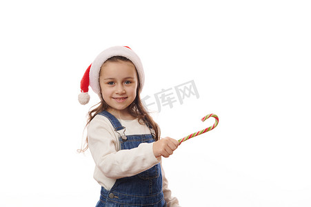 可爱的孩子，戴着圣诞帽的顽皮小女孩，用绿色的红色糖果手杖指着白色背景的复制空间