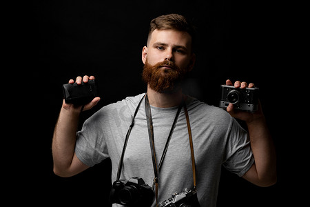 留着胡子的专业摄影师手里和肩上拿着一堆不同的老式旧相机。