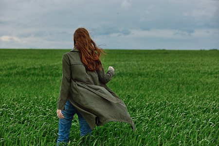 一位身穿长雨衣的红发女子在多云的天气中站在绿地里，在强风中