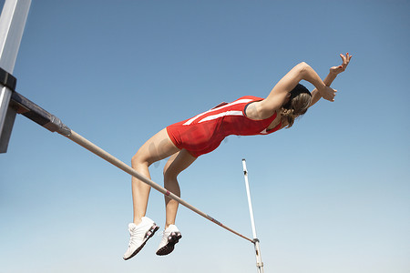 跳高运动员摄影照片_一名女子跳高运动员在半空中在蓝天的酒吧上空的低视角