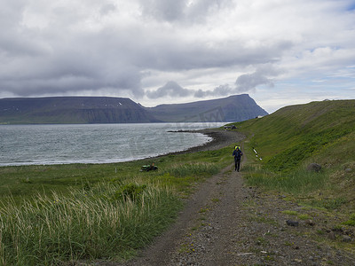 冰岛、西峡湾、Hornstrandir、Latrar，2018 年 6 月 26 日：背着沉重背包的孤独男子徒步者在自然保护区 Hornstrandir 的阿达尔维克湾的人行道上行走，那里有夏季小屋、绿草草甸、石滩、海洋、积雪覆盖的悬崖和
