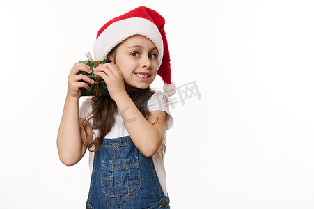 定位孩子摄影照片_戴着圣诞帽的淘气小女孩，摇着圣诞礼物，猜测里面是什么。