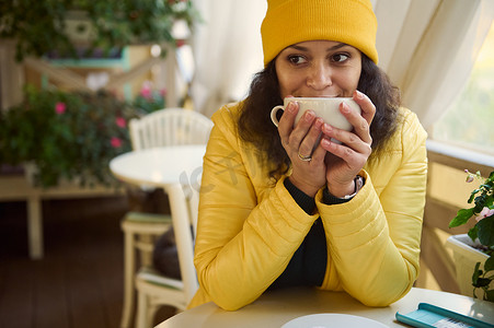 暖咖啡摄影照片_穿着黄色夹克的迷人女人，用一杯热饮暖手，喝一口咖啡，在咖啡店里放松