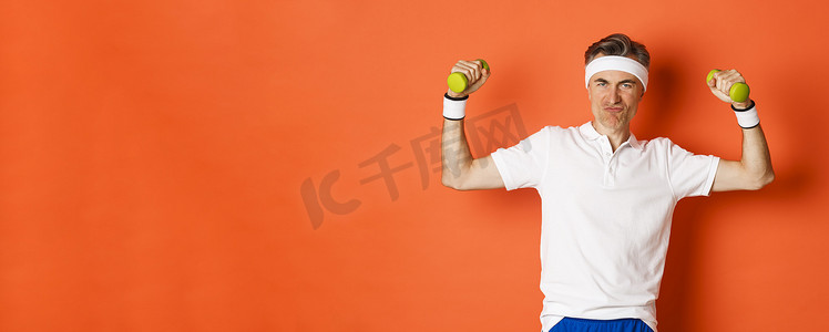 厚颜无耻的中年健身者的肖像，在橙色背景下做运动，弯曲二头肌并拿着哑铃