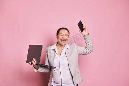 怀孕的中年女商人拿着笔记本电脑和手机，在粉红色背景中表达兴奋和喜悦