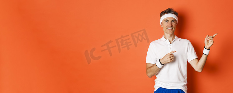 体育宣传背景摄影照片_身着头带和白色 T 恤的帅气男运动员的肖像，指着右上角微笑，展示体育宣传，站在橙色背景上