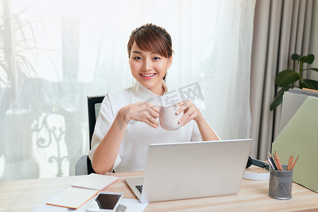在家用笔记本电脑工作时，美丽的亚洲年轻女性喝茶或咖啡