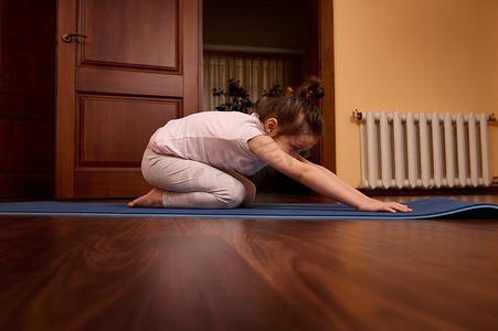 可爱的小女孩以儿童姿势伸展身体，在舒适的木制家居室内的蓝色健身垫上练习瑜伽