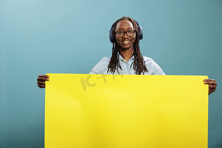 黄色广告海报摄影照片_戴着无线现代耳机的快乐积极女性手持黄色纸横幅标志