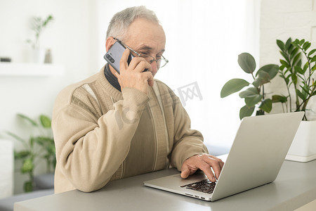 英俊微笑的老人戴着眼镜使用手机，坐在他舒适的工作场所，家里有笔记本电脑，退休男性在社交媒体上与朋友聊天，在智能手机上打字。