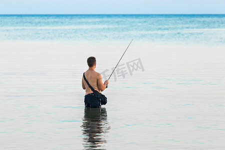 海水里的鱼摄影照片_一个没穿衬衫的小伙子晚上站在没膝的海水里，用鱼线钓鱼