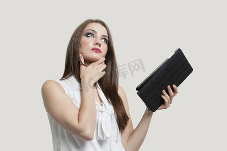 灰色背景下拿着平板电脑的沉思的年轻女人
