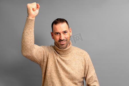 40 多岁留着胡须的西班牙裔男子身穿米色高领毛衣，举起拳头大力庆祝一场伟大的胜利，与世隔绝在灰色背景中