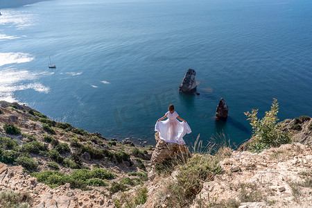 长腿摄影照片_在蓝天大海的映衬下，一位身穿白色轻裙长腿的美丽少妇挥舞着白色长裙站在海面之上的悬崖边上。