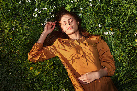 一个轻松的女人，躺在绿草地上休息，穿着橙色的长裙，闭着眼睛，脸上挂着愉快的微笑，正在休养