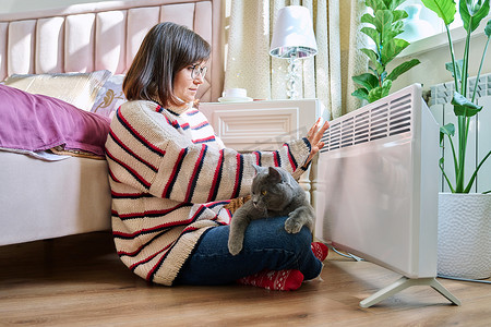 室内取暖摄影照片_女人和猫坐在一起，穿着温暖的毛衣，在电加热器旁取暖