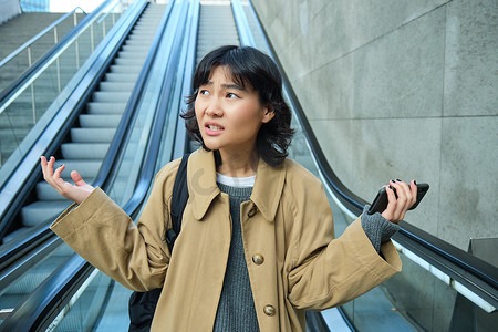 困惑的亚洲女孩的肖像不知道她在哪里，迷失在陌生的城市，带着沮丧的脸走下自动扶梯，耸耸肩，看着相机