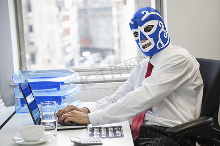 商人在办公桌上戴着摔跤面具使用笔记本电脑的肖像
