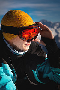 戴着滑雪护目镜的年轻滑雪者，里面反射着阳光，站在山上，把手放在头上，看着远方，肖像