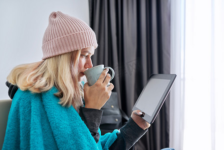 茶字摄影照片_戴着数字平板电脑茶杯的青少年学生坐在家里温暖的毯子下戴着帽子