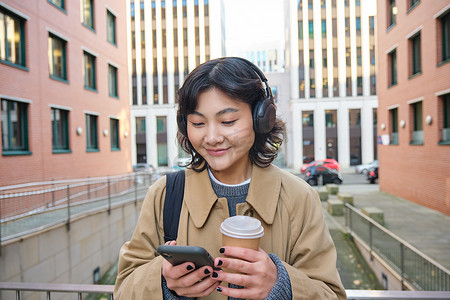 手机旅行app摄影照片_年轻女性，戴耳机的游客看智能手机，喝咖啡去，检查手机应用程序，听音乐和旅行