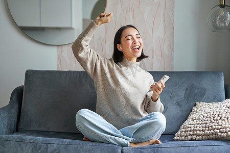 无忧无虑的亚洲女性在智能手机应用程序中唱歌和听音乐的肖像，使用无线耳机，微笑着高兴，坐在家里的沙发上