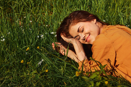 红色长围裙摄影照片_一个长着红色长发的平静女人躺在开着黄色花朵的绿色田野里，穿着橙色的裙子，愉快地微笑着，在灿烂的夏日阳光下闭上了眼睛