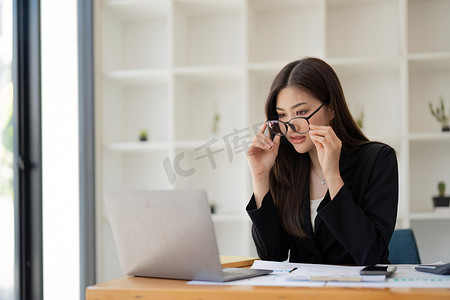 坐在办公桌前的漂亮女商人拿着眼镜，周围是电脑笔记本电脑、文书工作、计算器、与财务、办公室税务相关的文件