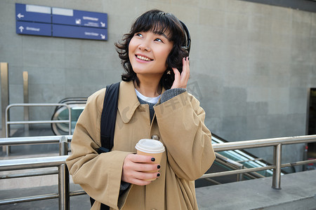 年轻学生的画像，戴耳机的女孩，喝咖啡，背着背包站在街上，上大学或学院，开心地微笑