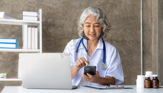 医生办公室摄影照片_身穿白色医用大衣的白发女医生正在使用智能手机，坐在办公室时微笑着使用笔记本电脑