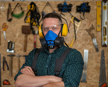 呼吸器摄影照片_在车间里，一位戴着护目镜、耳机和呼吸器的木匠双臂交叉在胸前站立。