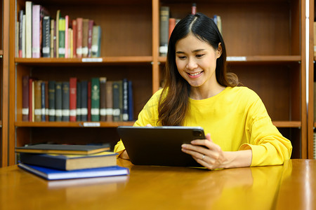 微笑的女大学生准备考试，在图书馆的数字平板电脑上搜索信息