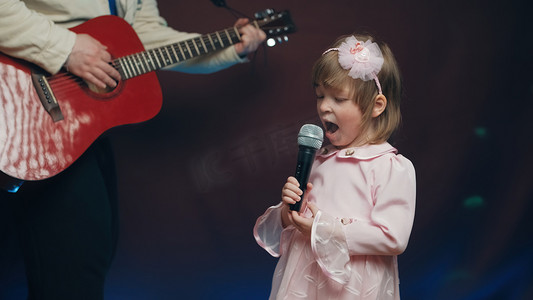儿童节音乐摄影照片_穿着复古裙子的小女孩在舞台上唱歌，她的父亲弹着原声吉他