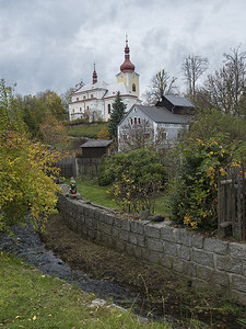 在 Luzicke hory 的 Sloup v cechach 村的教堂和房屋的景色，Lusatian 山脉有秋天的彩色树木和小水流，蓝天，白云