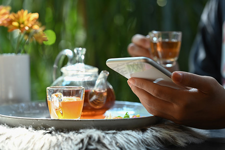 在质朴的木桌上用杯子和茶壶在传统茶具附近使用智能手机的女性近景