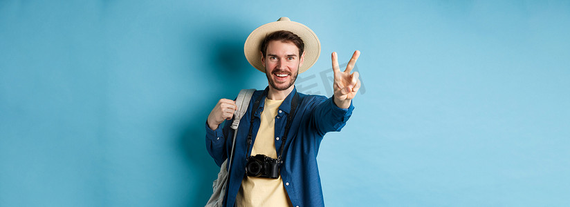 戴草帽的男摄影照片_帅气积极的男游客在暑假展示和平标志，在阳光下享受热带假期，戴着草帽，拿着背包和相机，蓝色背景