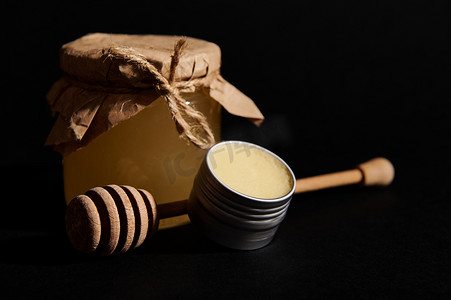 玻璃瓶中有机蜂蜜的静物生活，罐头上有蜡和木制蜂蜜勺，突显在黑色背景中