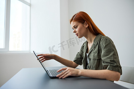 红头发的女人坐在办公室靠窗的桌子旁全职工作，在笔记本电脑上打字