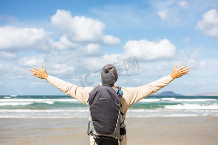 在西班牙兰萨罗特岛 Famara 多风的沙滩上，年轻的父亲在背包里背着他的婴儿男婴儿子享受纯净的大自然，同时举起双手向天空。