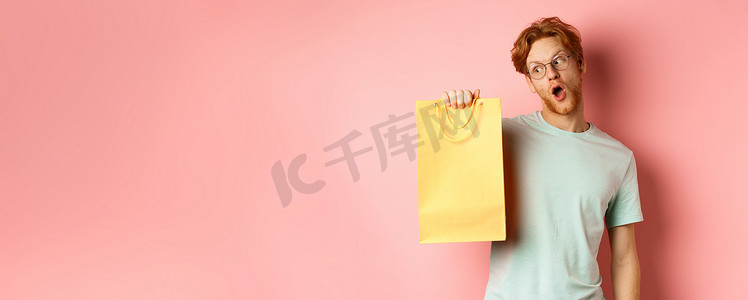 红头发、戴着眼镜和 T 恤、拿着并看着黄色购物袋、在促销活动中购买礼物、站在粉红色背景上的风趣帅哥