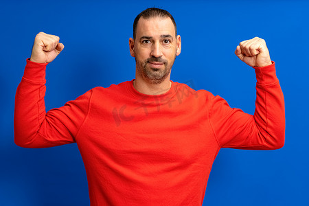 40 多岁的西班牙裔男性快乐、不刮胡子、强壮的运动健身学生穿着红色时髦毛衣，手上显示二头肌肌肉，与蓝色背景工作室肖像隔离。