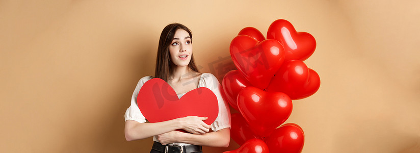 大红色浪漫摄影照片_浪漫的女孩拥抱情人节大红色的心形剪纸，站在浪漫的气球旁边，向左看标志，梦幻般地凝视着促销，米色背景